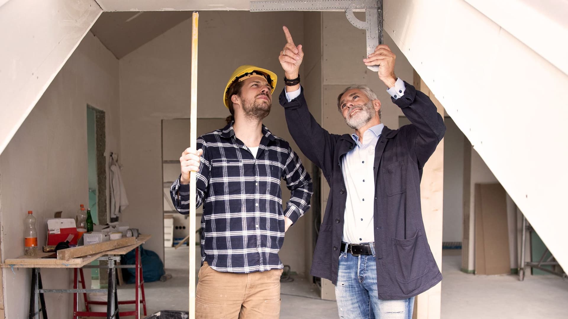 Zwei Männer arbeiten gemeinsam an einem Dachbodenausbau, während sie Materialien und Werkzeuge organisieren. Der Raum ist hell erleuchtet und zeigt eine Mischung aus Holzrahmen und Dämmmaterialien, die auf den bevorstehenden Umbau hinweisen.