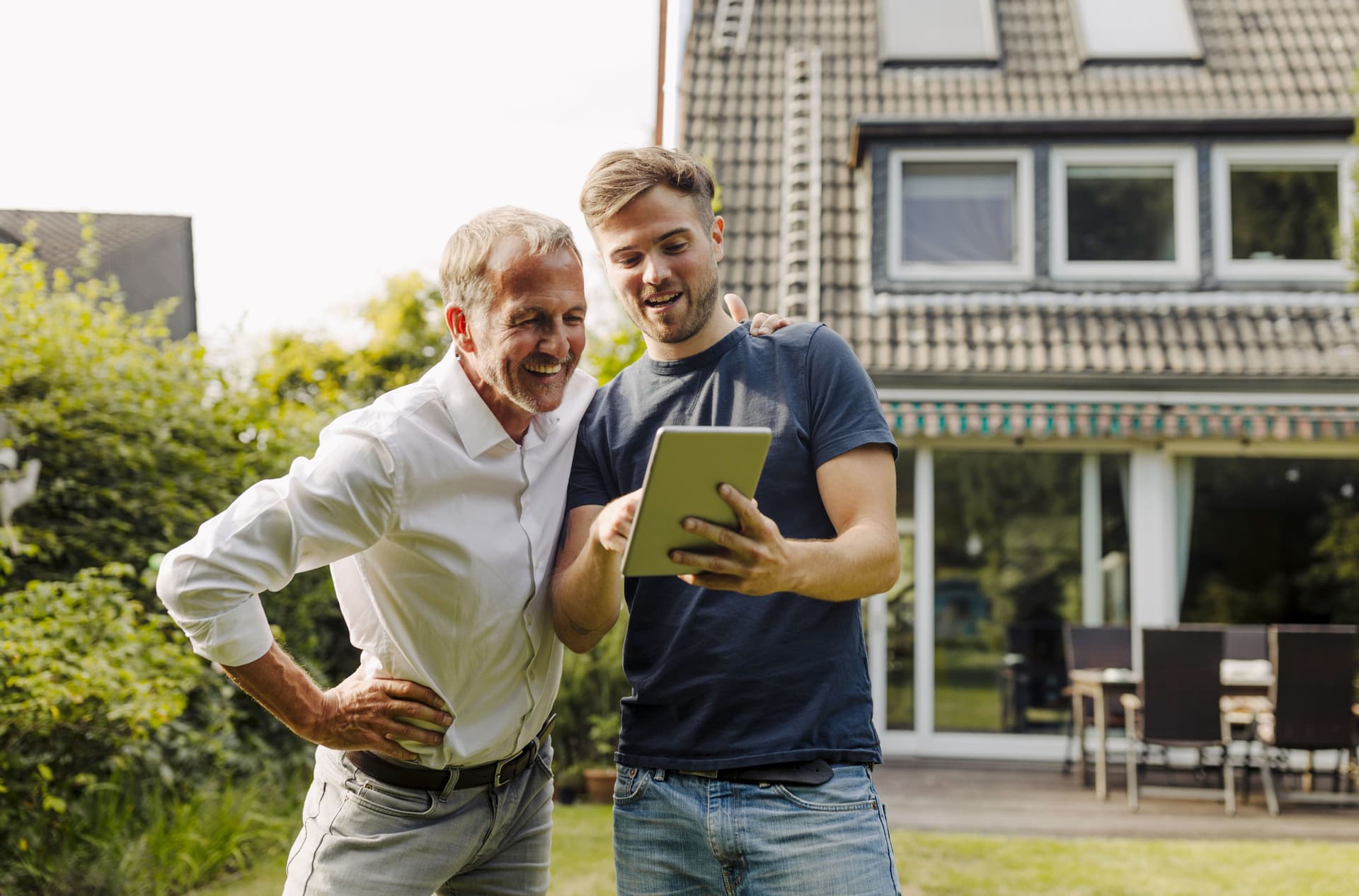 Zwei Männer stehen im Garten mit einem Tablet in der Hand. Sie schauen auf das Tablet. Im Hintergrund ist ein Einfamilienhaus zu sehen.