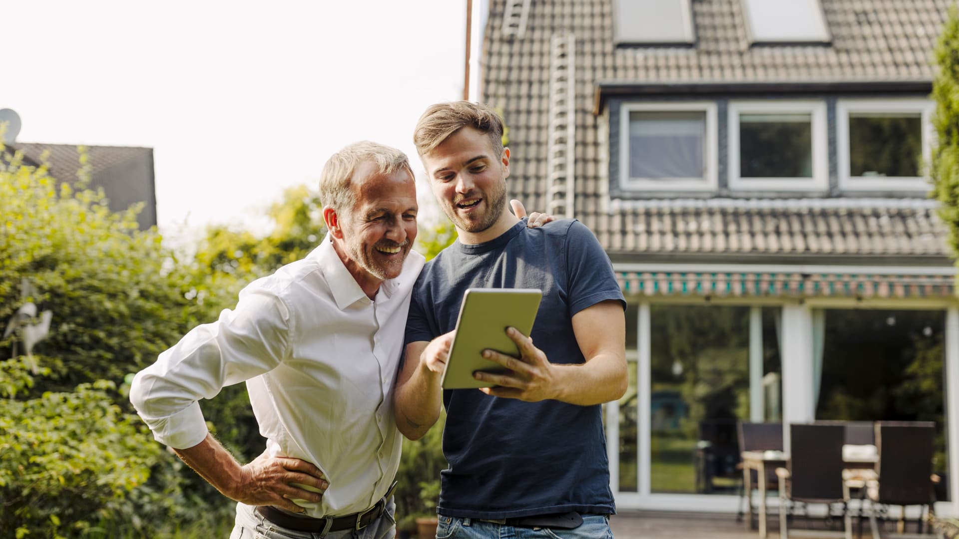 Zwei Männer stehen im Garten mit einem Tablet in der Hand. Sie schauen auf das Tablet. Im Hintergrund ist ein Einfamilienhaus zu sehen.